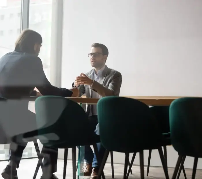 Dos hombres se sientan frente a frente en una mesa de despacho y discuten.